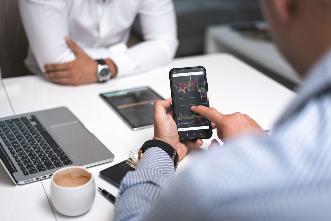 Offerings Two businessmen analyze stock market data using a smartphone and laptop in a modern office setting.