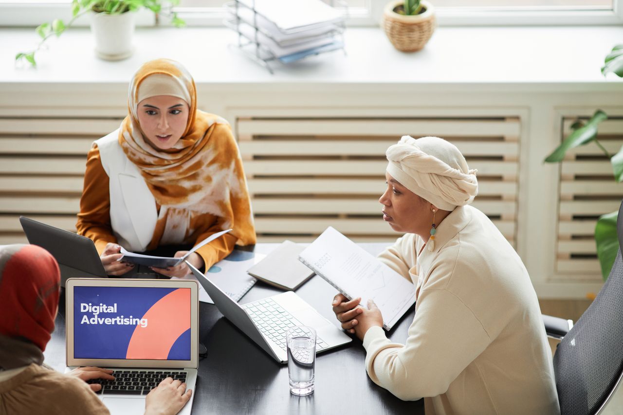 Home Three women in hijabs collaborating on digital advertising strategies around a table with laptops.